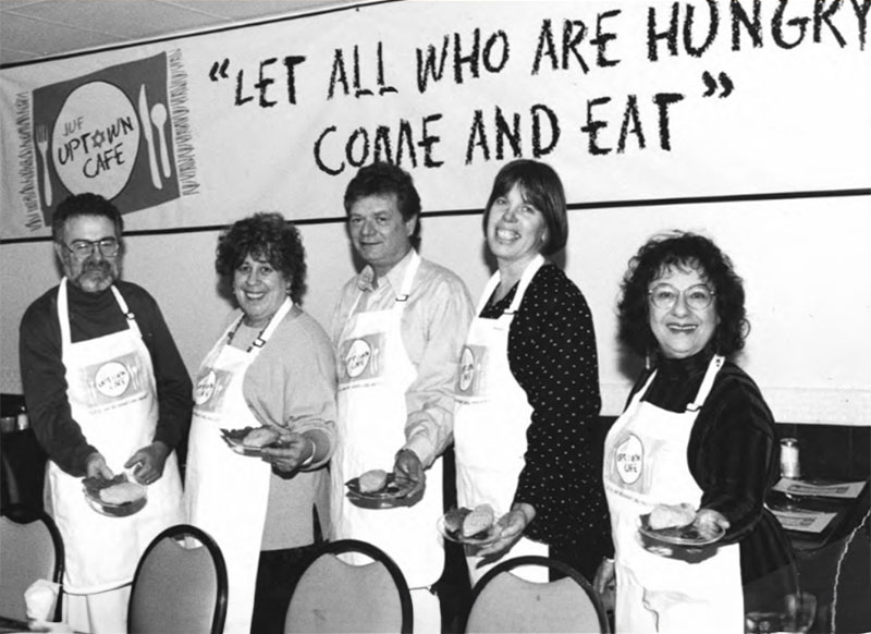 A black and white photo of five Ezra volunteers holding out plates of food with a sign behind them reading "Let all who are hungry come and eat."