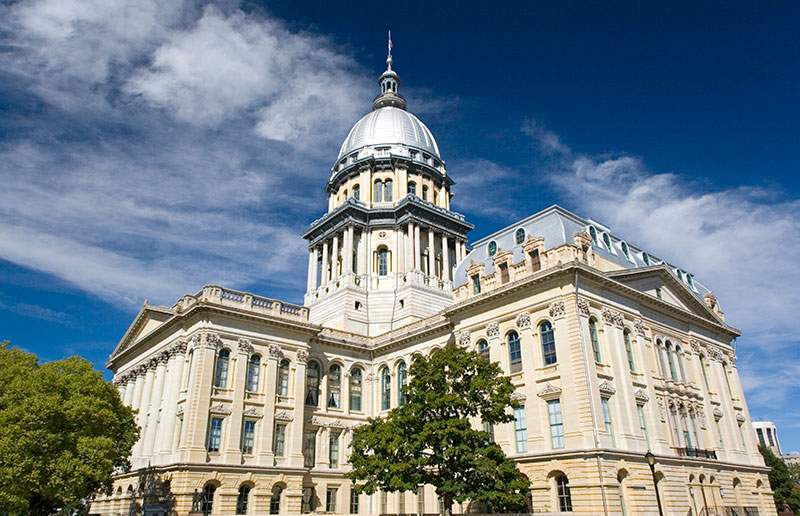 Illinois State capitol with green trees, blue sky with white clouds in the background. The building is cream with a tall grey dome and pointed tip with American flag at the top.