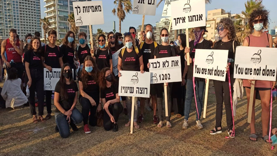 A group of individuals standing on a beach in Tel Aviv wearing matching shirts holding signs that read "you are not alone".