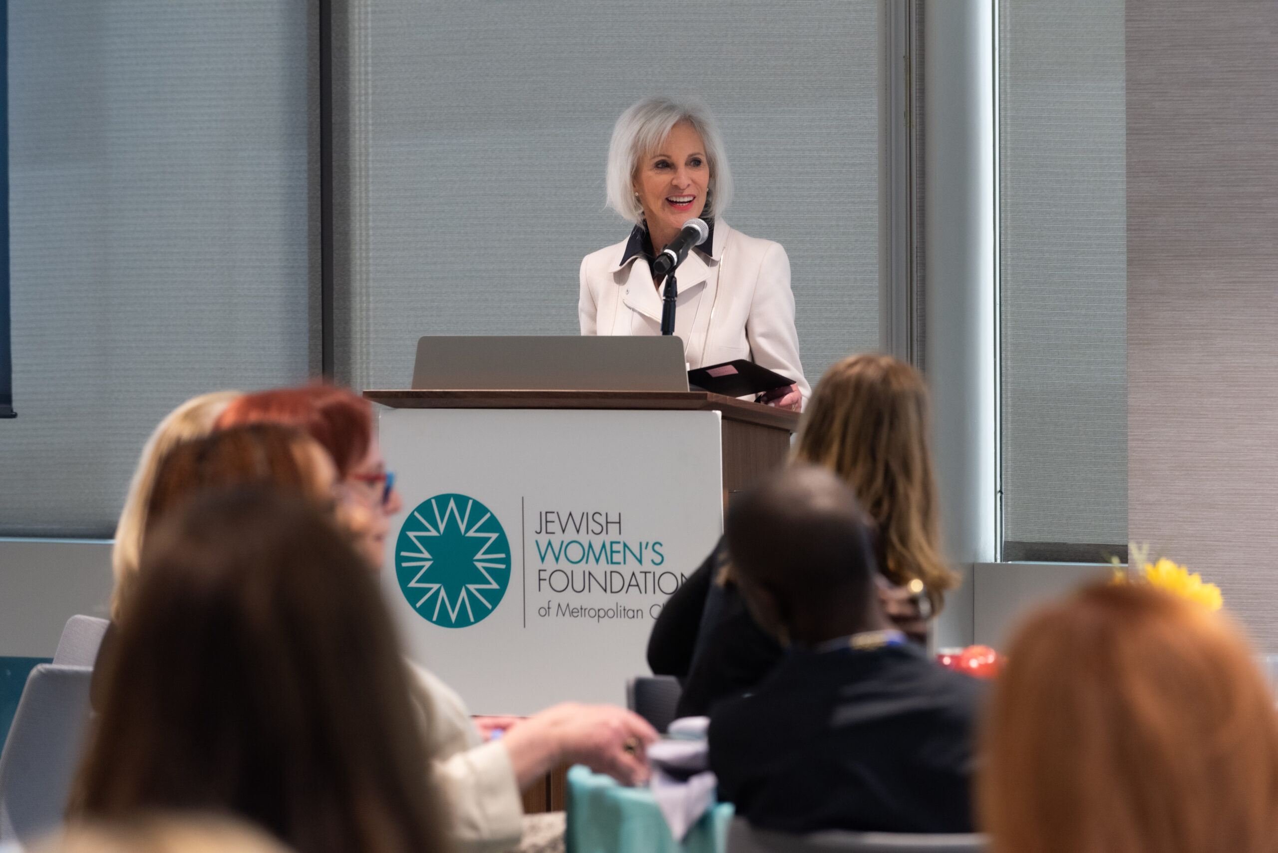 A woman speaking at a podium featuring the "Jewish Women's Foundation of Metropolitan Chicago" logo.