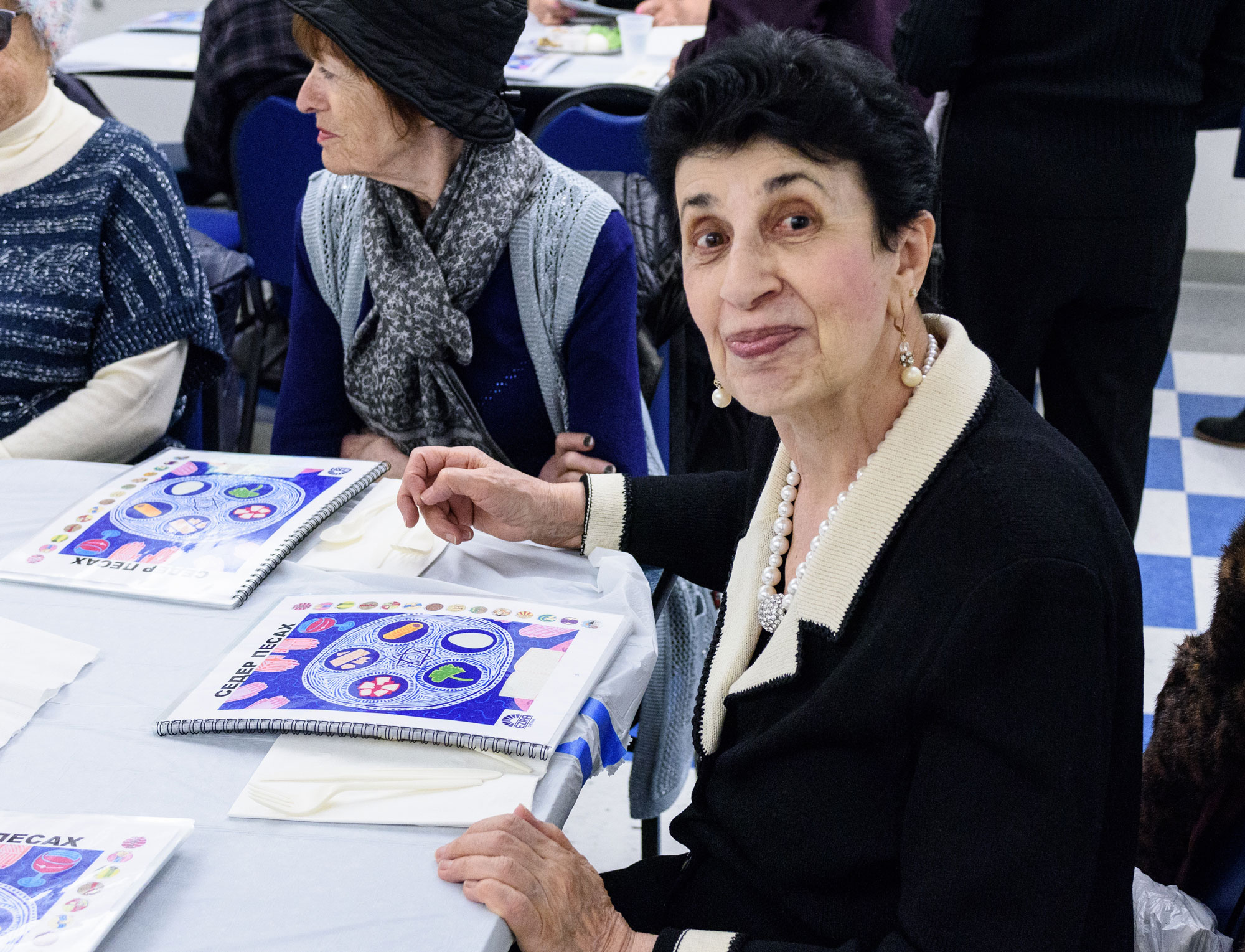 An older Jewish woman with short black hair wearing pearls reading a Russian seder book for Passover.