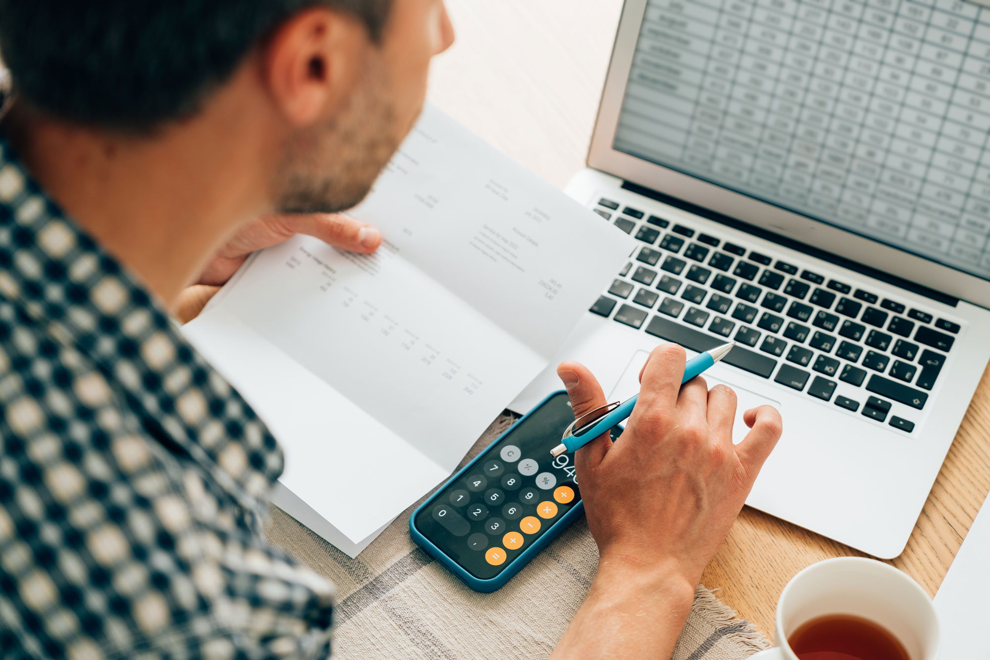 Close up photo from above angle showcasing a man typing on a MacBook with his iPhone calculator app open and a pen and paper.