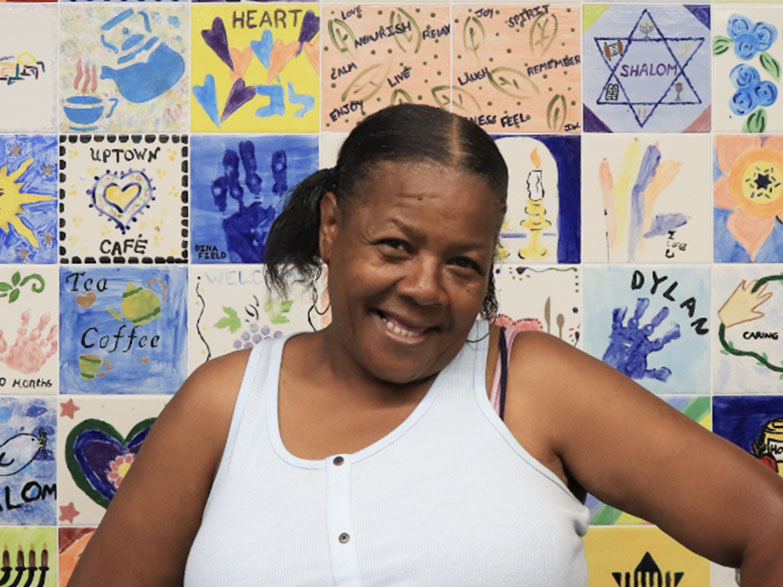 A woman in a white tank top smiling and posing in front of a tile mural.