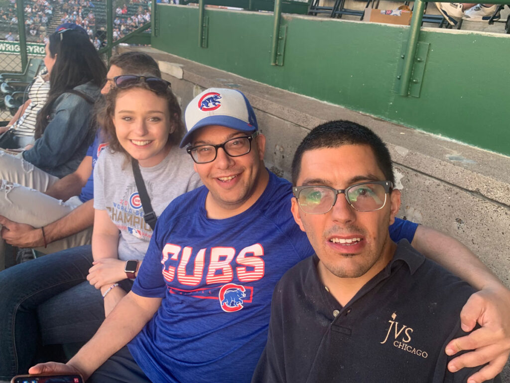 Two men with disabilities and a woman all wearing glasses sitting at a cubs game smiling for a selfie.