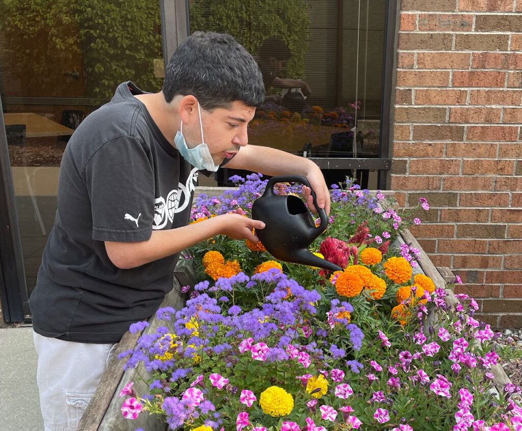 A man with disabilities using a black watering can to water a flower bed on a porch.