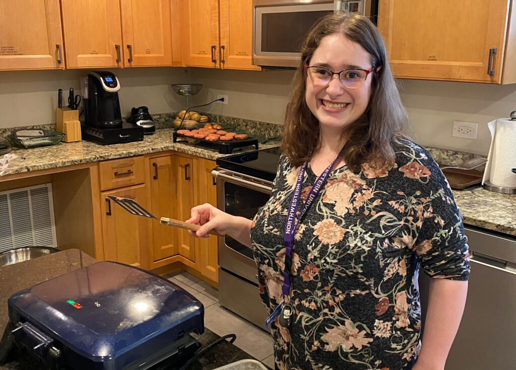 A woman in a floral shirt with a lanyard holding a spatula smiling while cooking in the kitchen.