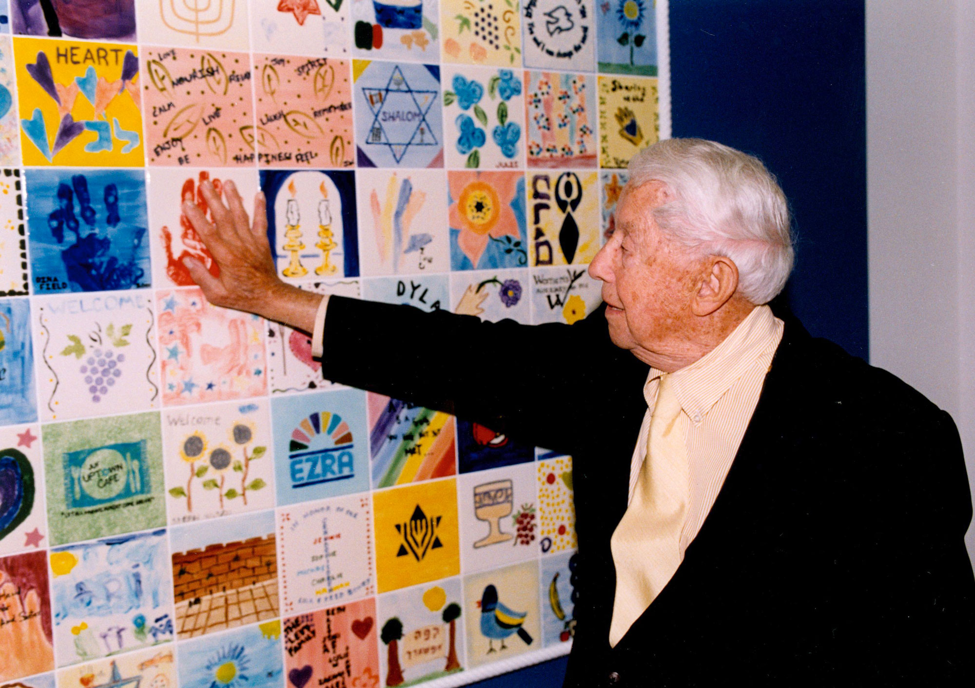 An old man in a suit putting his palm up on a mural wall of Ezra creations.