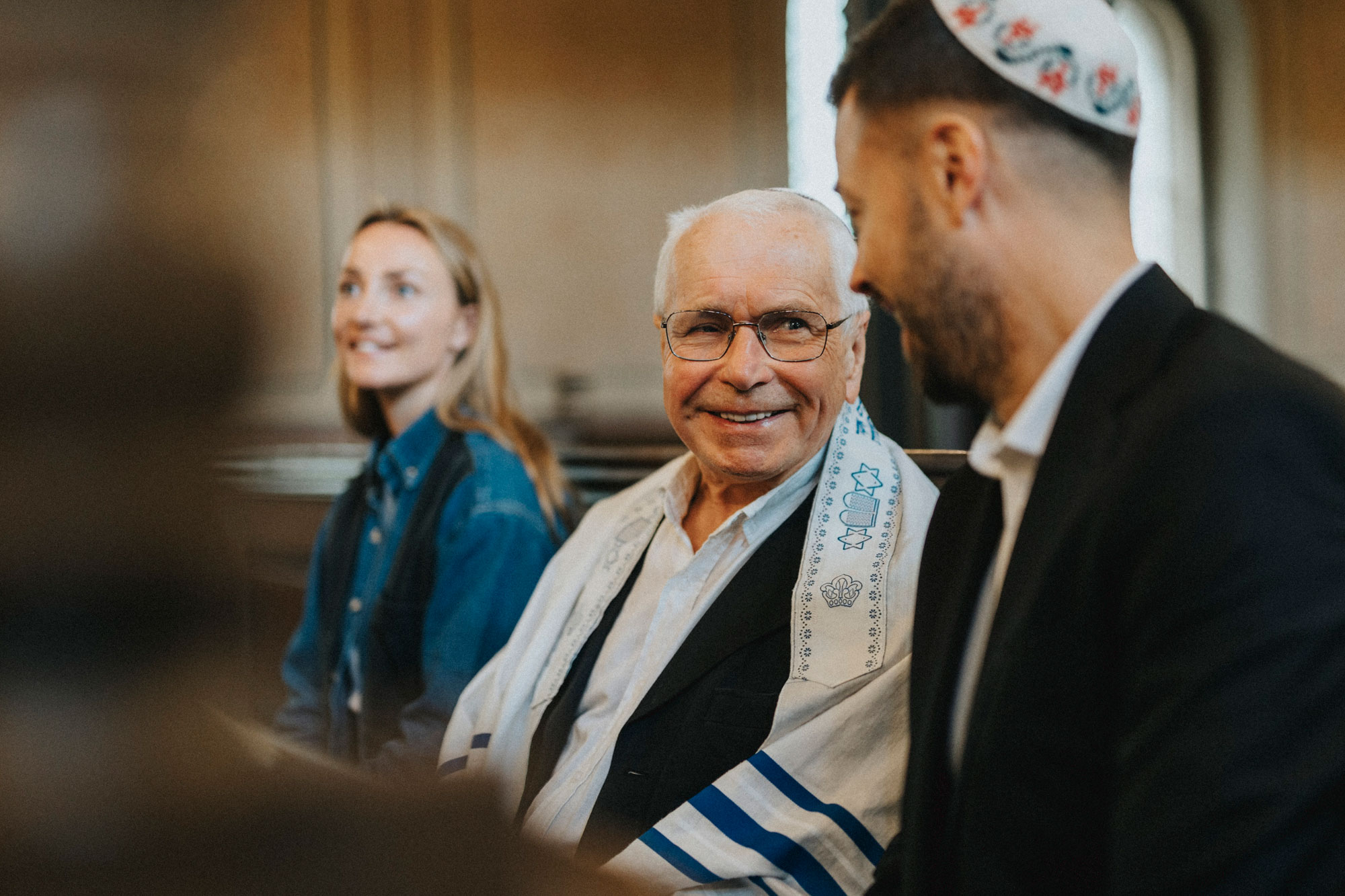Jewish synagogue with three individuals, one wearing a tallit and kippah, indoors with natural light.