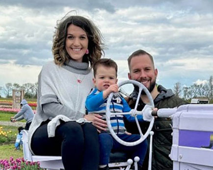 Jeanette Pacanowski and sitting on a tractor in a garden with her young son using the steering wheel beside Jeanette's husband.