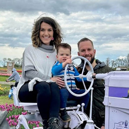 Jeanette Pacanowski and sitting on a tractor in a garden with her young son using the steering wheel beside Jeanette's husband.