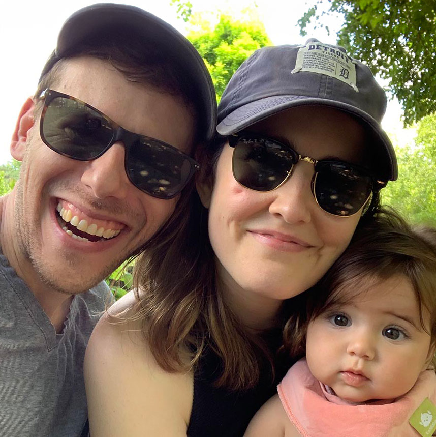 Kelsey Rosenfeld wearing smiling in a selfie with her husband and baby girl. Kelsey and her husband are both wearing glasses and hats.