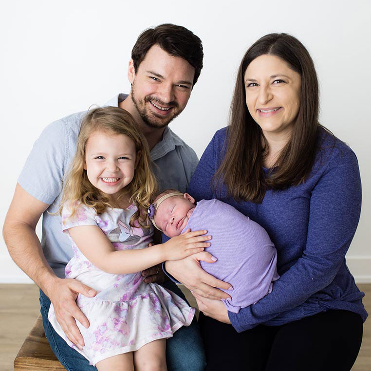 Kimberly Schulte and her husband smiling for a portrait with their toddler daughter smiling and newborn infant both on their laps while all wearing various shades of purple.