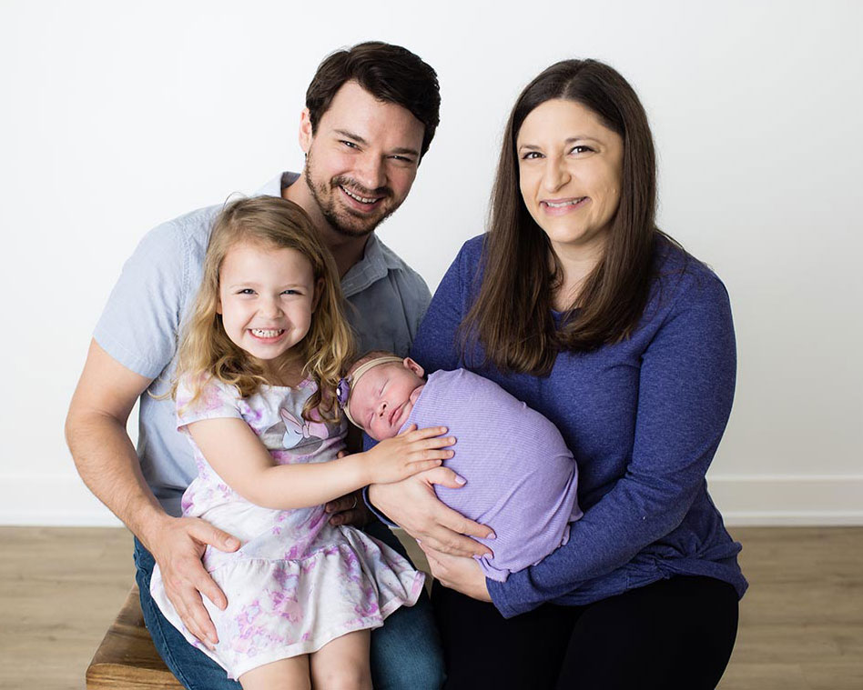 Kimberly Schulte and her husband smiling for a portrait with their toddler daughter smiling and newborn infant both on their laps while all wearing various shades of purple.