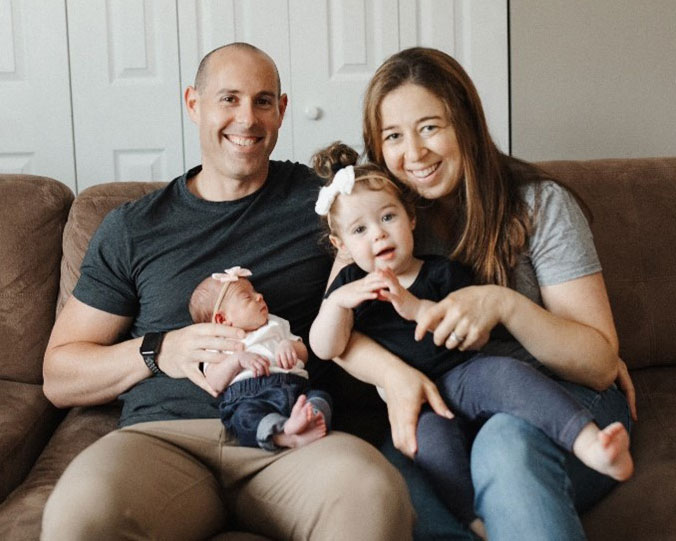 Lauren Koenig and her husband sitting on a couch smiling while holding their newborn baby and their young daughter both wearing pink bows on their heads.