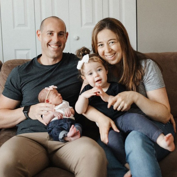 Lauren Koenig and her husband sitting on a couch smiling while holding their newborn baby and their young daughter both wearing pink bows on their heads.