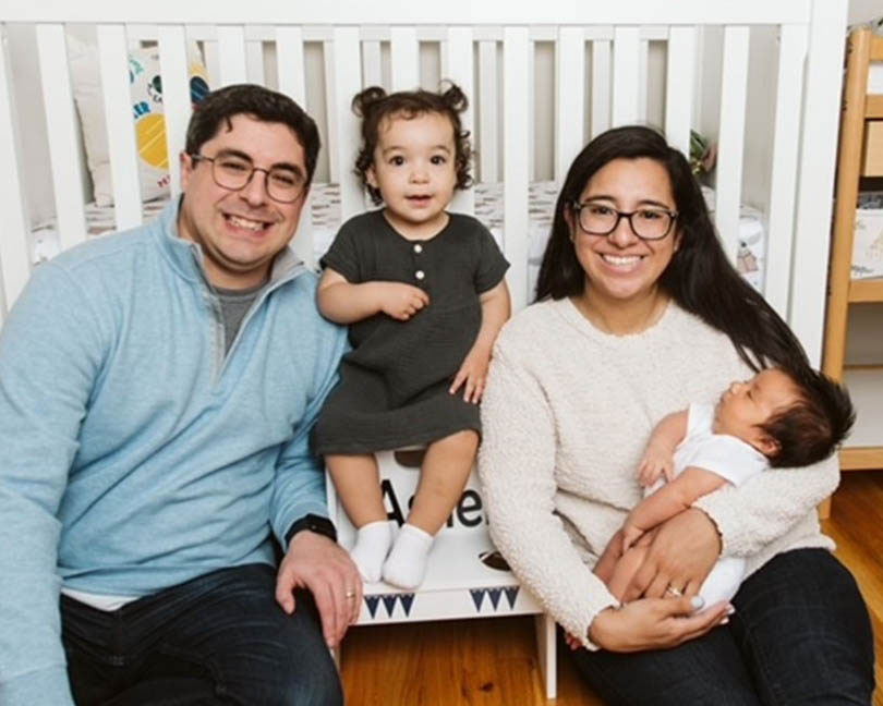 Leah Venus and her husband smiling in glasses on the floor in front of a crib with their young toddler daughter and their new born baby in arms.