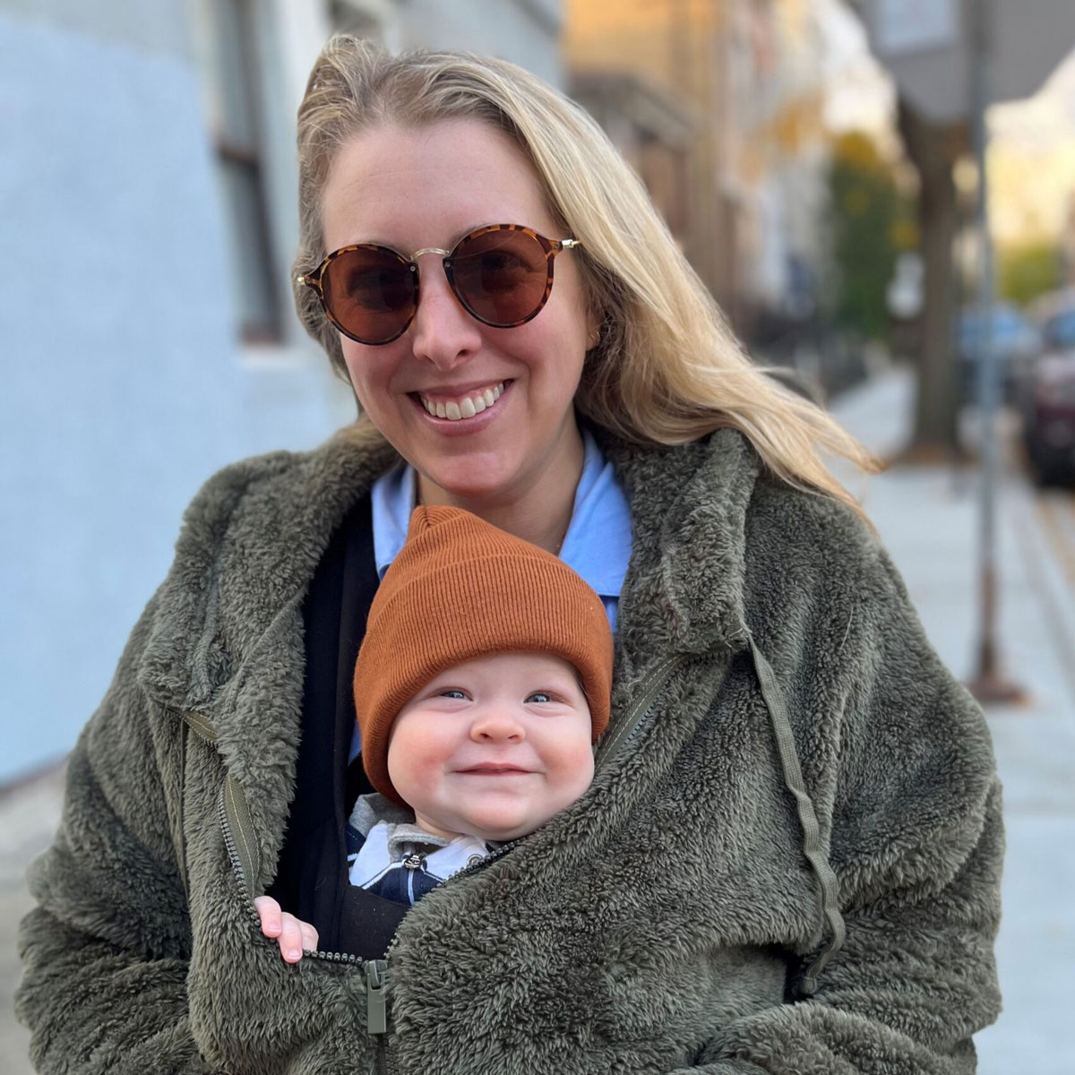 Marissa Rhines holding her young baby on her chest both smiling at the camera. The baby is wearing a beanie and Marissa is wearing sunglasses.