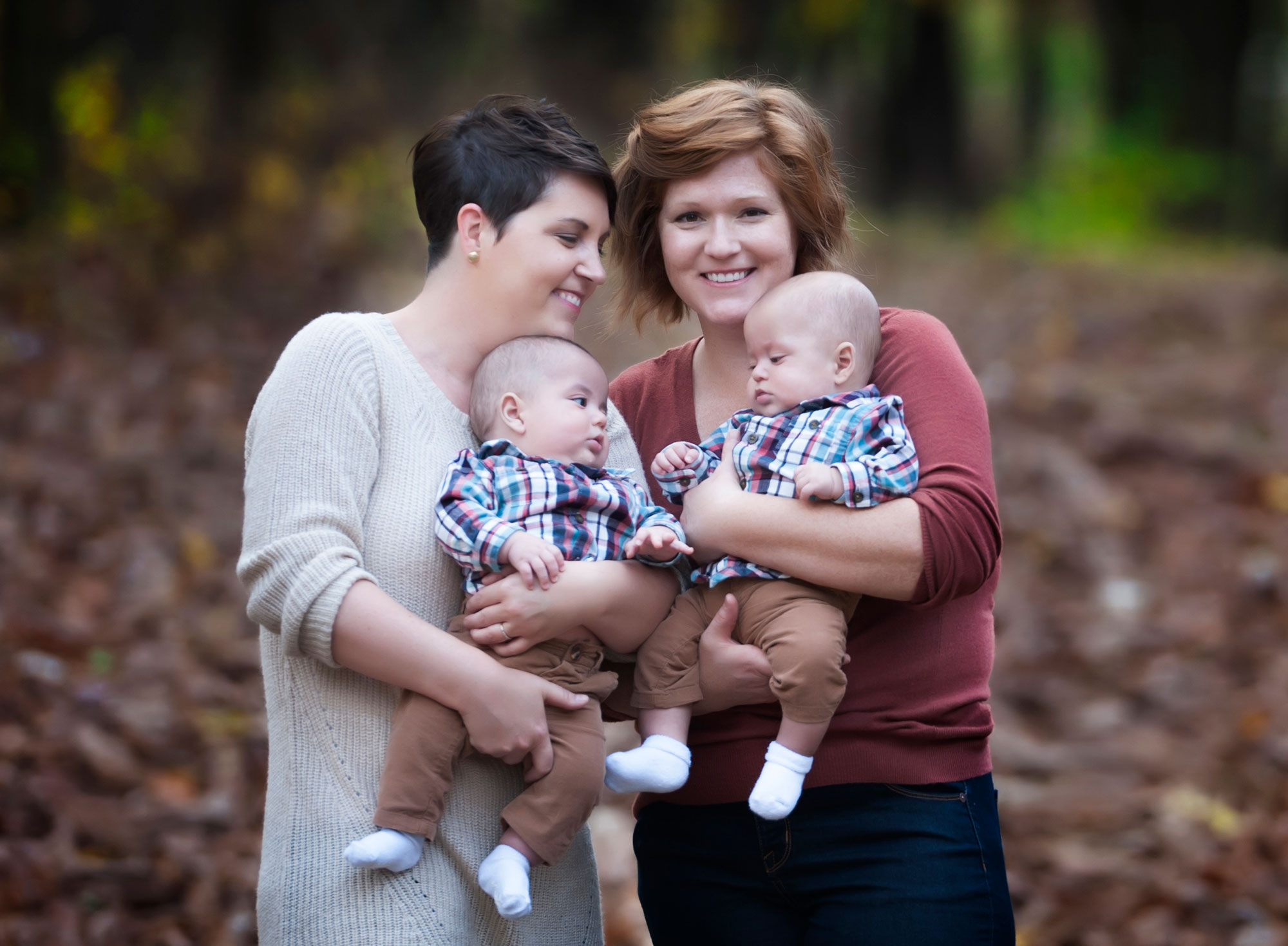 Two women with short hair holding twin babies wearing identical outfits including plaid shirts with brown pants with a fall leafy blurred background.