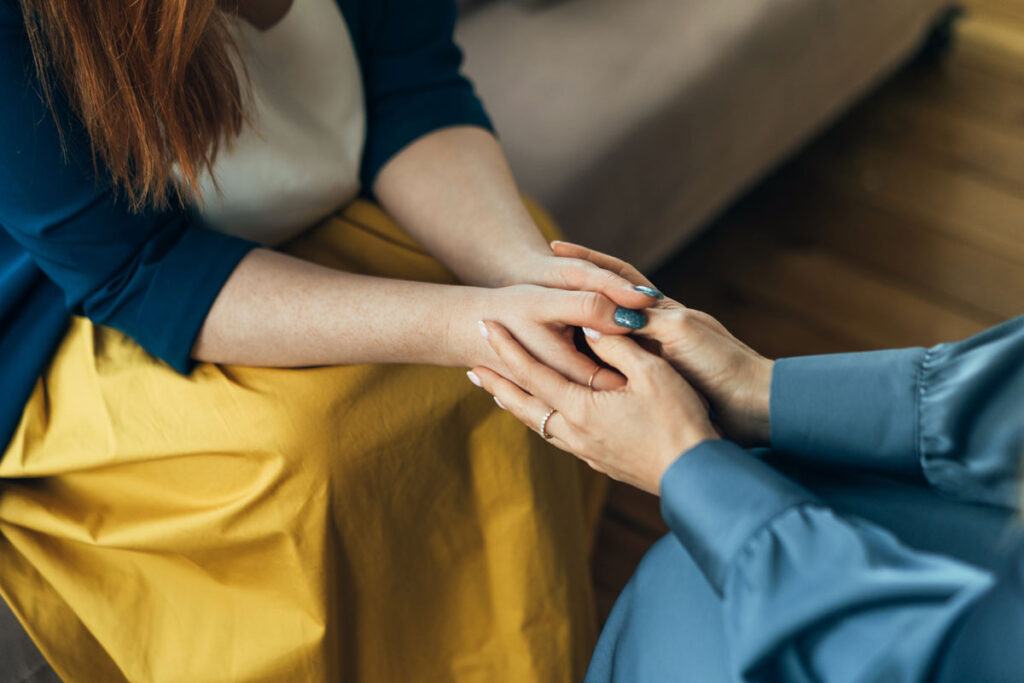 A zoom in of one woman holding another woman's both hands. The women are both wearing rings and their nails are painted.