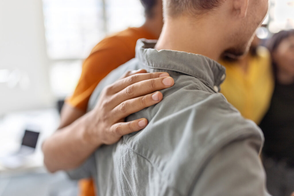A closeup of a hand placed on a man's shoulder who is wearing a gray collared shirt. No faces are shown.