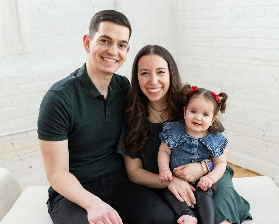 Shmira Barnett and her husband sitting on a ledge with a white background holding their young baby daughter who is wearing two red bows in her hair and they are all smiling in a portrait.