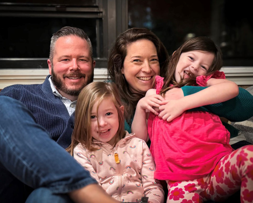 Talia Treece and her husband sitting on the couch holding their two young daughters all smiling.