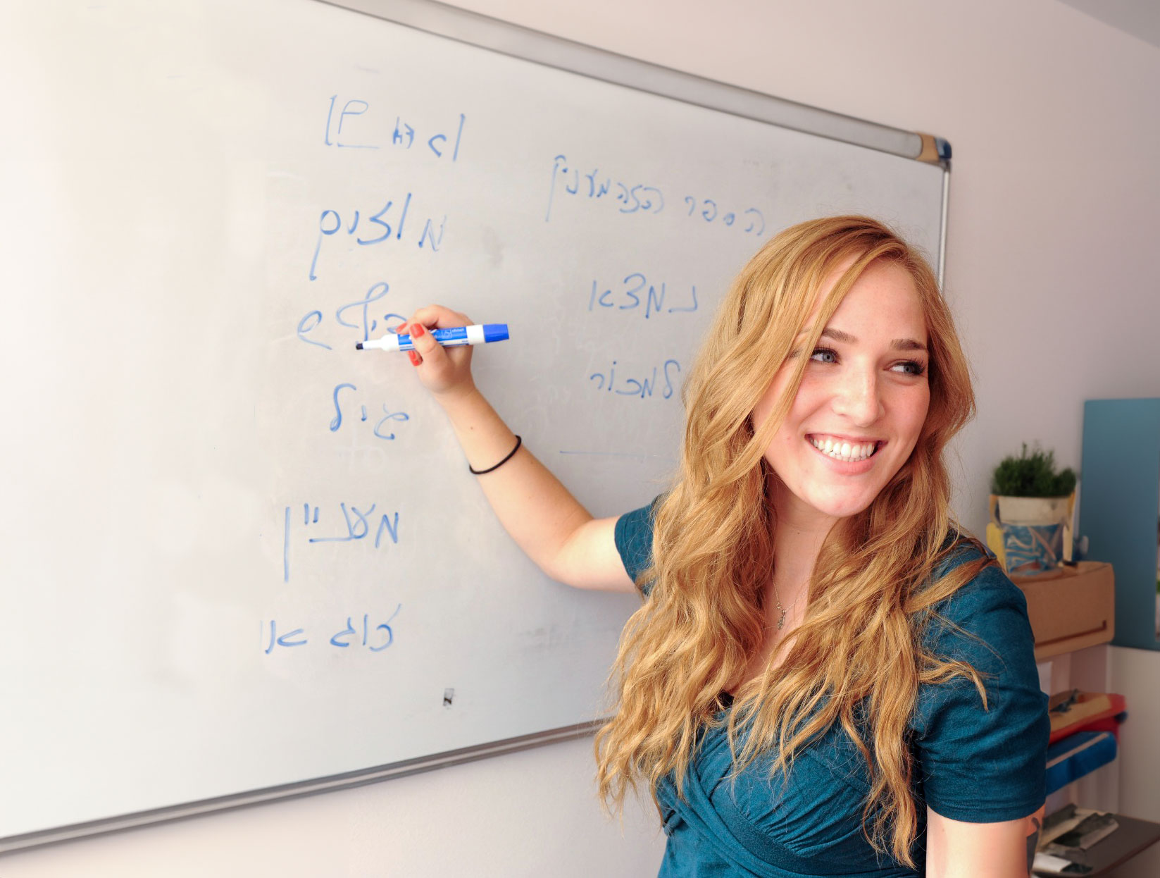 A blond woman smiling while writing in Hebrew in blue expo marker on a dry erase board.
