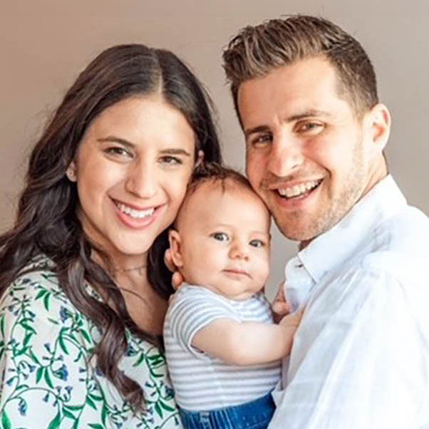 Whitney Ash Zucker and her husband smiling while holding their baby in the middle of them in a portrait photo.