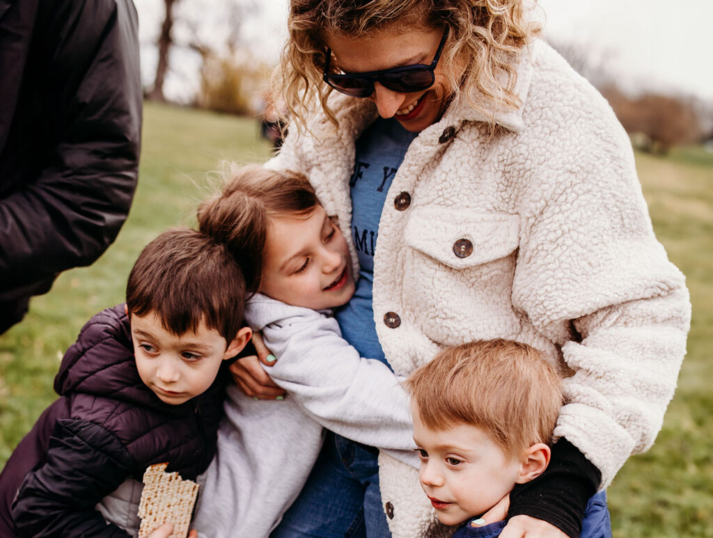 A mom wearing sunglasses looking down at her three kids all group hugging her.