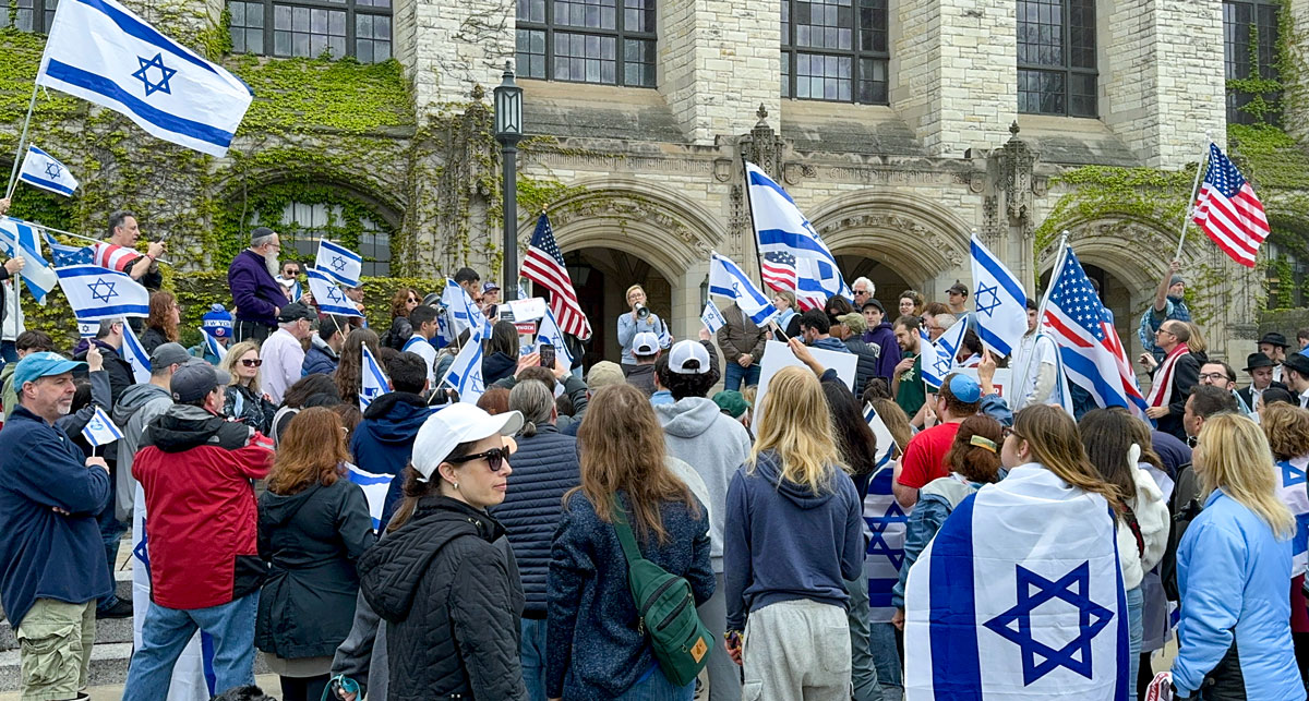 A crowd of people waving Israeli flags listening to a speaker with a megaphone on a college campus.