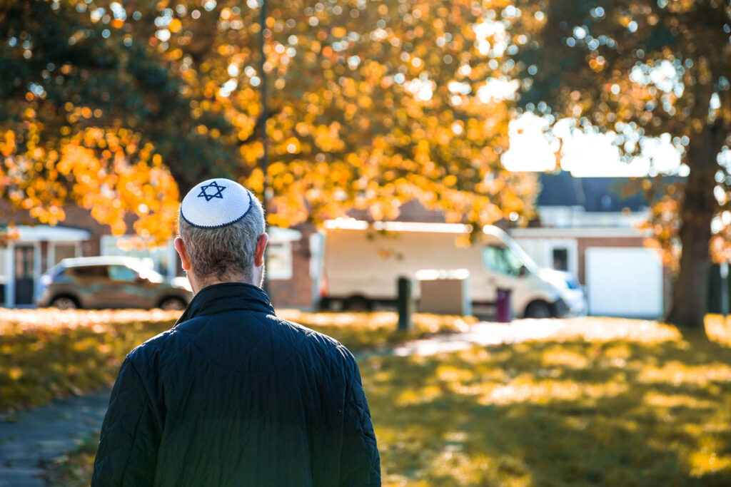 The back of a man wearing a white kippah with a blue Jewish star facing an autumn neighborhood.