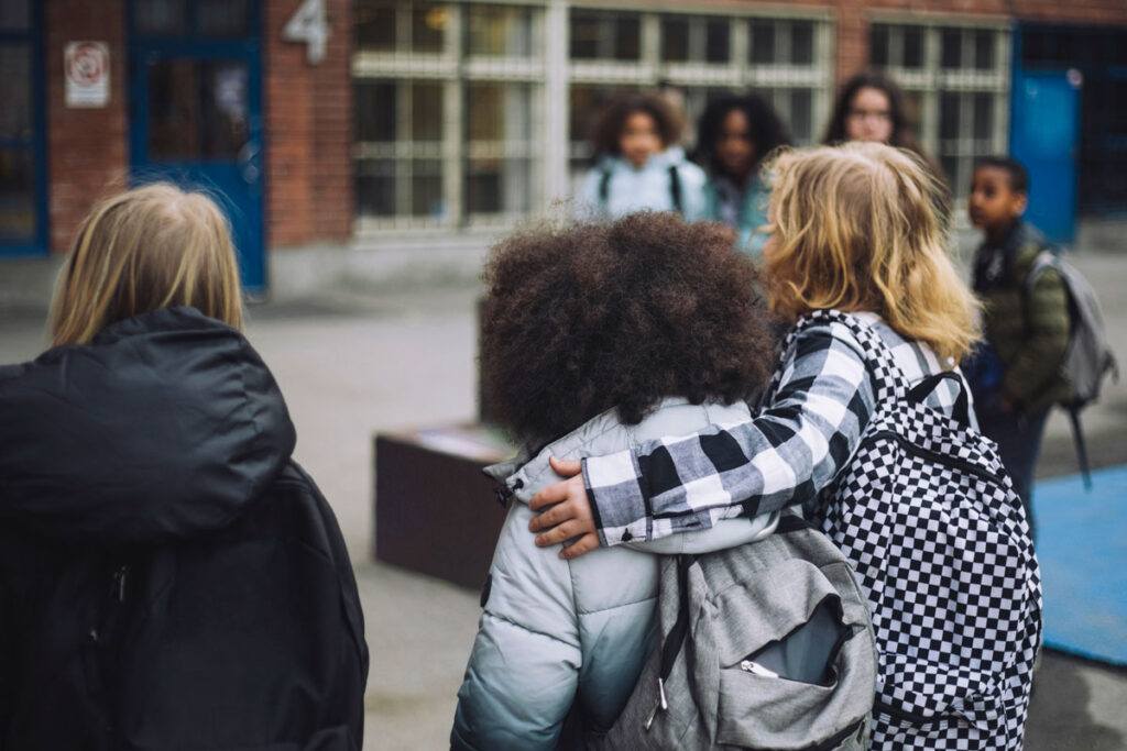 Two college students wearing backpacks with arms around each other standing on campus.