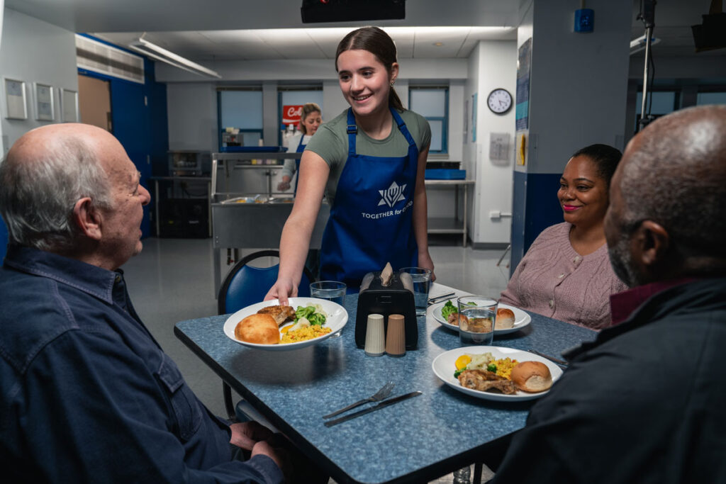 A girl wearing a blue JUF "Together for Good" apron serving people plates of food at a dining hall.