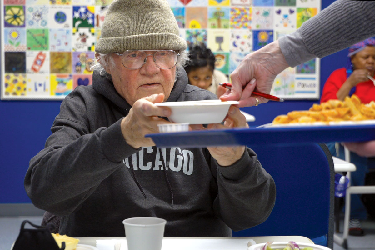 An older man wearing a green beanie and a "Chicago" sweatshirt receiving soup at the EZRA cafe.