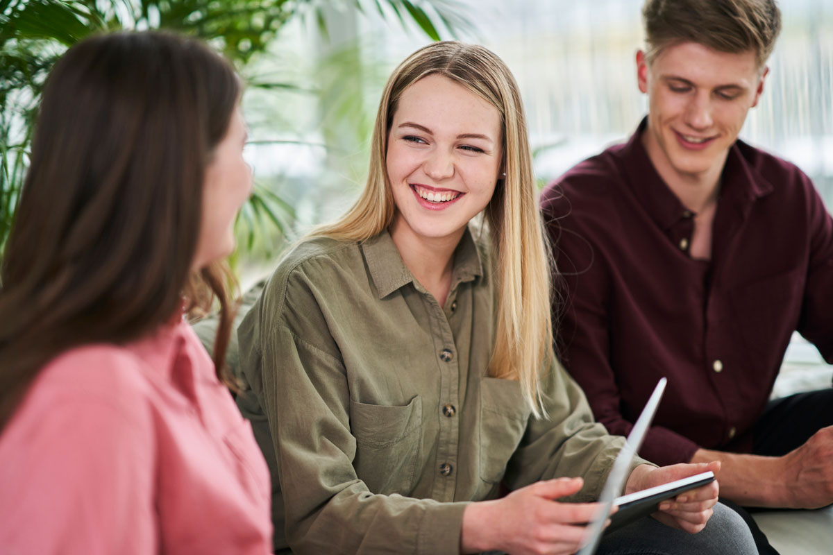 Three college students sitting with notebooks featuring one blonde woman in the middle wearing a green blouse.
