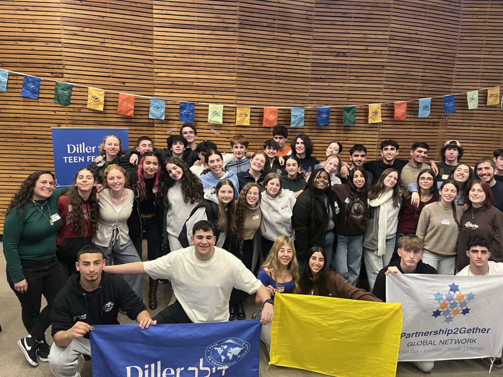 A large group of participants gathered indoors, smiling and holding banners for Diller Teen Fellows and Partnership2Gether, standing in front of colorful hanging flags.