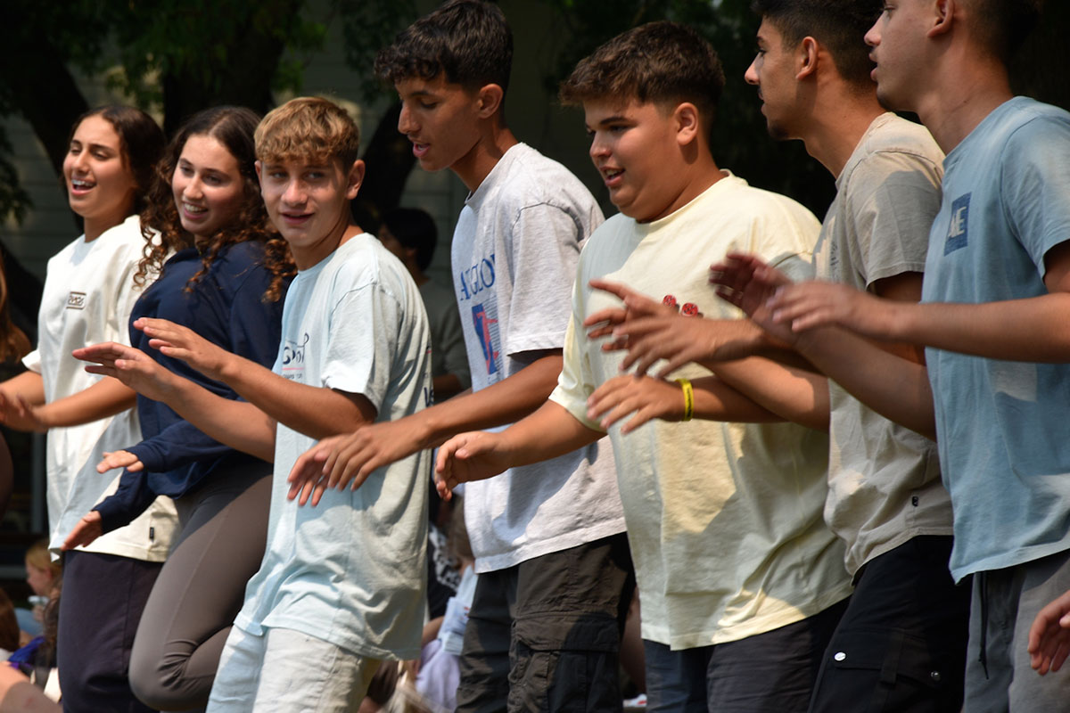 A group of Nir Oz teens standing in a row outdoors, moving their arms in unison as if participating in a coordinated group performance.