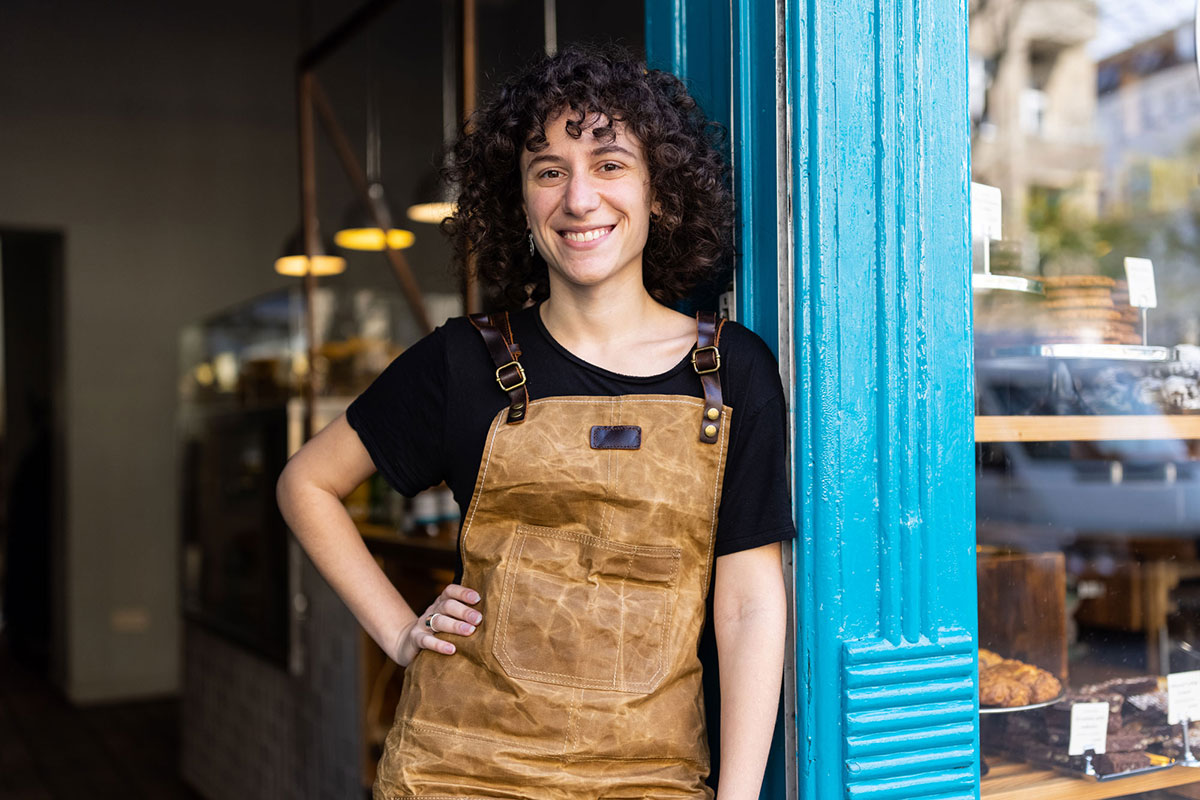 An individual wearing a brown apron leans against a bright blue storefront, standing near a bakery display with pastries visible through the window.