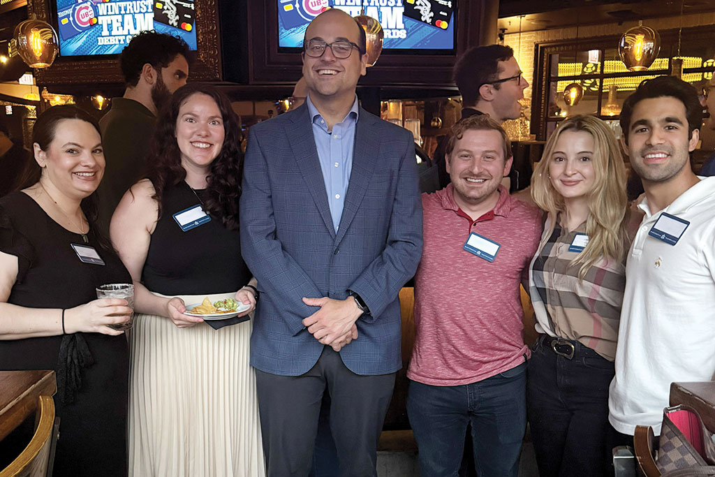 Group of professional men and women standing together at a social event with TV screens displaying graphics in the background.