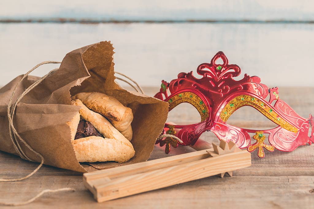 Hamantaschen in a brown paper bag next to a pink Purim mask and a wooden grogger.