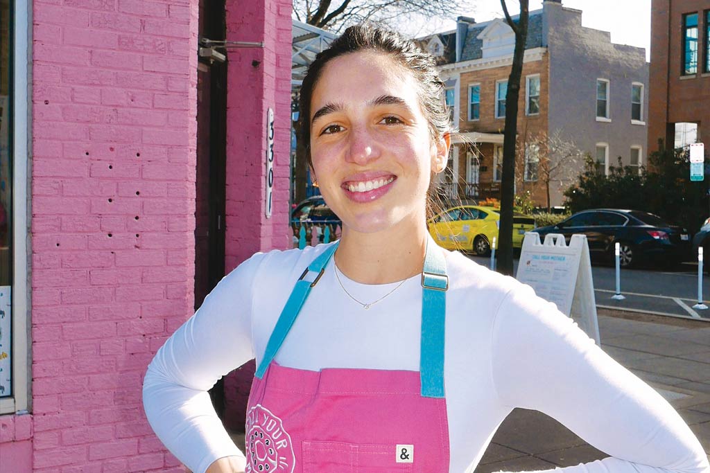 Daniela Moreira wearing a white long sleeve shirt and pink apron outside her bagel shop.
