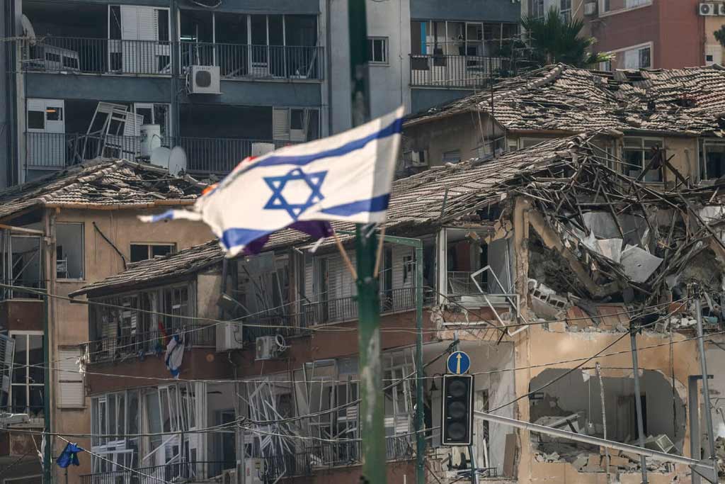 Israeli flag flying in the wind in font of houses in rubble after Iranian missile attack.
