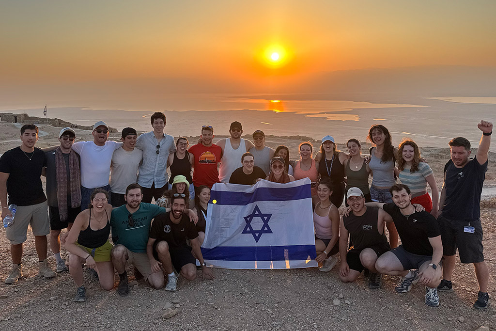 A group of teens holding an Israeli flag with a sunset in the background